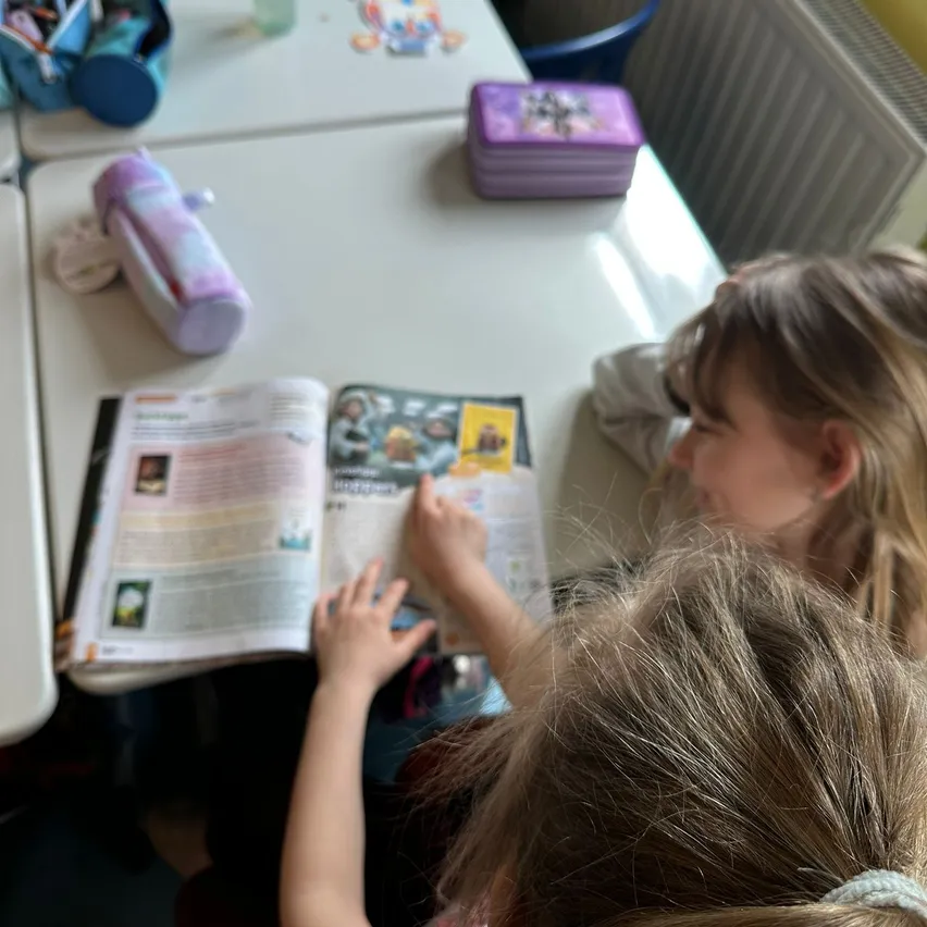 Zwei junge Mädchen sitzen an einem Schreibtisch und lesen ein Buch. Eine Tasche und andere Gegenstände liegen auf dem Tisch. Ein Heizkörper ist an der Wand hinter ihnen angebracht.
