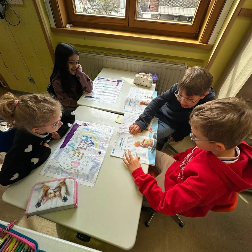 Vier Kinder sitzen an einem Tisch und beschäftigen sich mit einer Malaktivität. Zwei Jungen und zwei Mädchen, ein Mädchen lächelt, während die anderen sich auf ihre Malbücher konzentrieren.