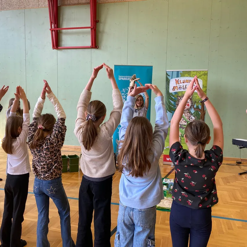 Eine Gruppe junger Mädchen in einer Turnhalle, die Yoga-Posen üben. Sie haben ihre Hände über den Kopf erhoben. Im Hintergrund ein blauer Banner mit weißem Text.