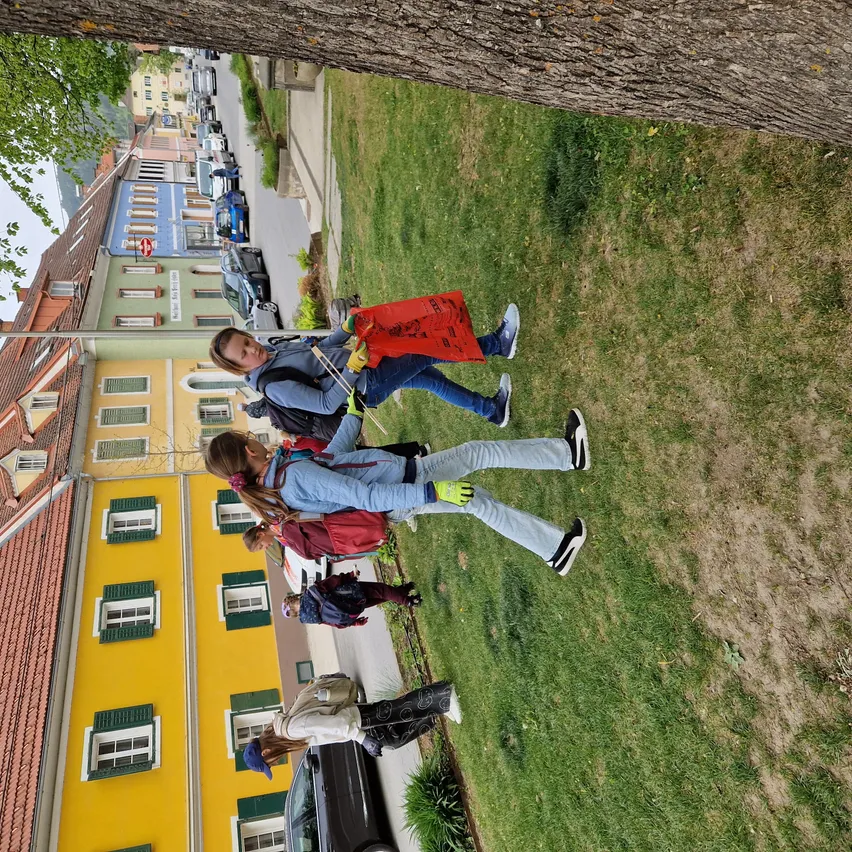 Eine Gruppe von Menschen sammelt Müll auf einer Straße. Sie tragen Handschuhe und halten rote Beutel. Die Straße ist von geparkten Autos und Gebäuden gesäumt. Ein Gebäude hat eine gelbe Wand mit grünen Fensterläden.