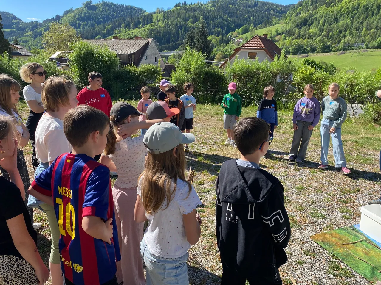 Eine Gruppe von Kindern ist auf einem Rasen versammelt. Einige tragen Mützen und Brillen. Sie scheinen jemandem zuzuhören. Der Bereich hat einige Pflanzen, und Häuser sind im Hintergrund.