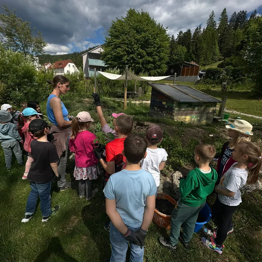 Eine Gruppe von Kindern mit einer Frau pflanzen in einem Garten mit einem Schuppen und einem bewölkten Himmel im Hintergrund.