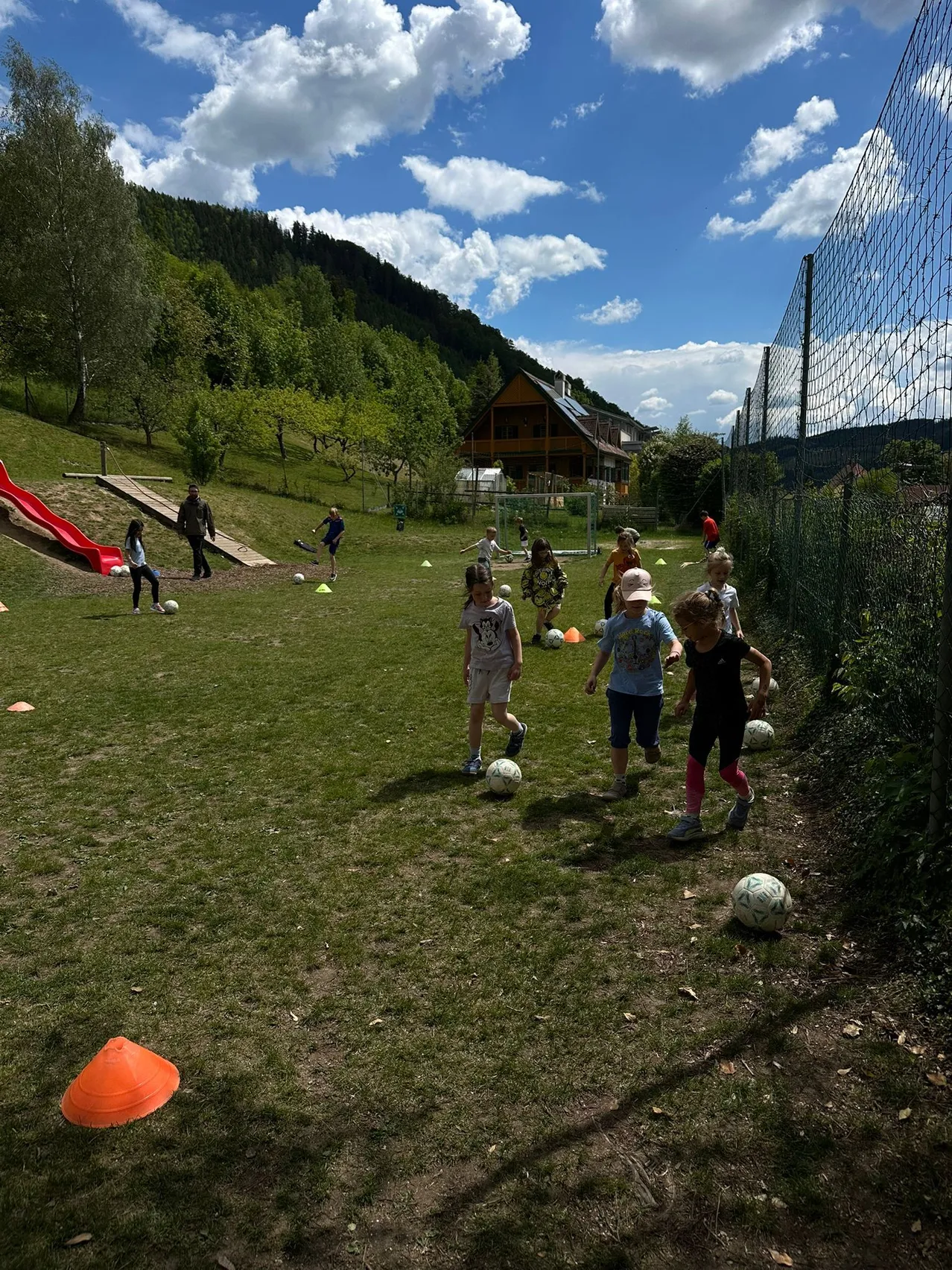 Eine Gruppe von Kindern spielt Fußball auf einem Rasenfeld mit orangefarbenen Kegeln. Ein roter Rutsch und ein Haus sind im Hintergrund.