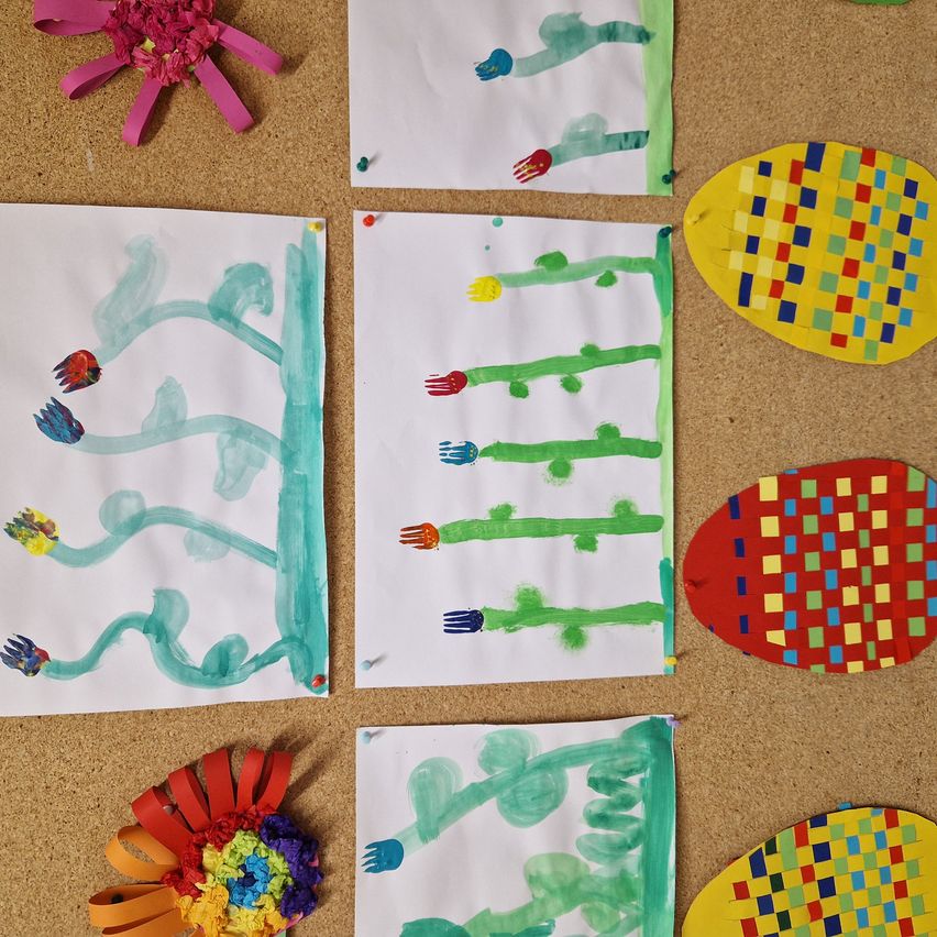 Mehrere Kunstwerke sind auf einer braunen Oberfläche ausgelegt. Vier weiße Blätter mit blauen und grünen Zeichnungen sind ausgestellt. Es gibt auch drei bunte, quadratisch gemusterte Kunstwerke. Zwei von ihnen sind rot und gelb, und eines ist blau und grün.