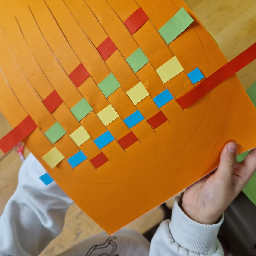 Eine Person hält ein orangefarbenes Papier mit einem gewebten Muster, bestehend aus verschiedenen farbigen Quadraten und Rechtecken, auf einer Holzoberfläche.