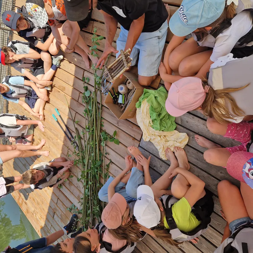 Eine Gruppe von Kindern sitzt im Kreis auf einer Holzplattform und schaut einen Mann an, der eine kleine Holzkiste in der Hand hält. Sie tragen Hüte und Rucksäcke.