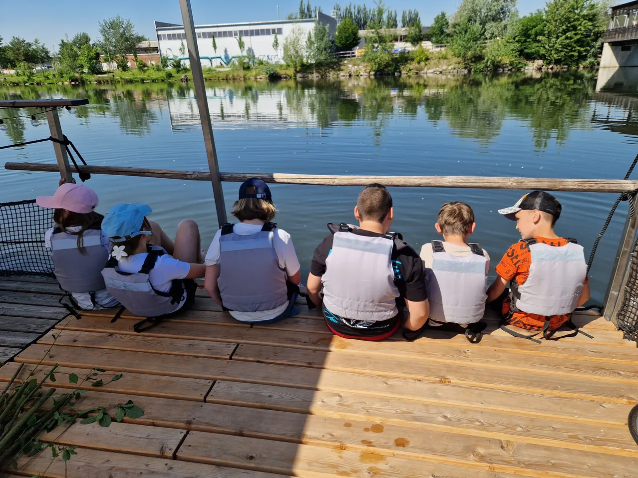 Fünf Kinder sitzen auf einem Holzsteg am Wasser. Sie tragen alle Schwimmwesten. Sie schauen etwas im Wasser an. Das Wasser ist ruhig. Es gibt Bäume und ein Gebäude in der Ferne.
