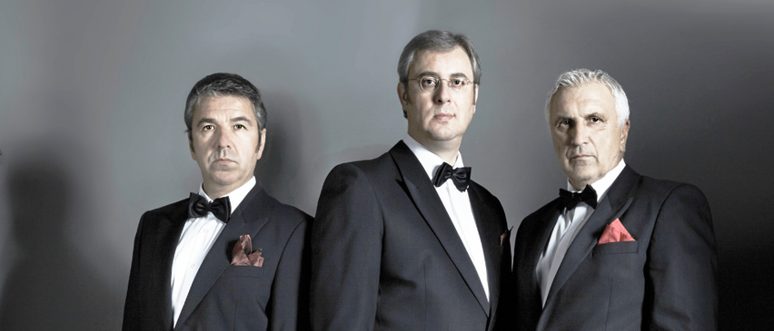 Drei Männer in formellen Anzügen stehen nebeneinander vor einem grauen Hintergrund. Der Mann auf der linken Seite trägt ein rotes Taschentuch, während der Mann in der Mitte eine Brille trägt. Der Mann auf der rechten Seite hat ein rotes Taschentuch in der Tasche.