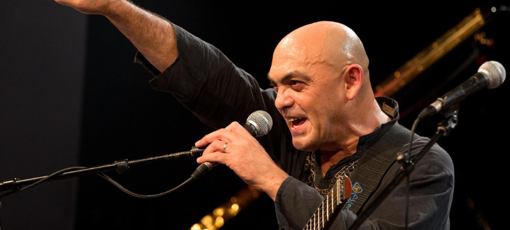 Ein kahlköpfiger Mann singt und spielt Gitarre auf der Bühne, hält ein Mikrofon und trägt ein schwarzes Hemd. Hinter ihm ist ein verschwommener Hintergrund eines Klaviers.