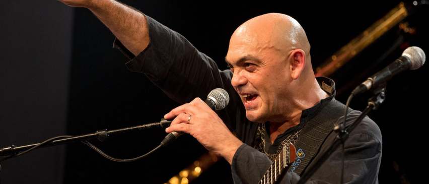 Ein kahlköpfiger Mann singt und spielt Gitarre auf der Bühne, hält ein Mikrofon und trägt ein schwarzes Hemd. Hinter ihm ist ein verschwommener Hintergrund eines Klaviers.