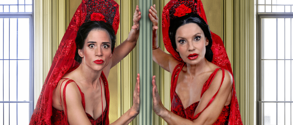 Zwei Frauen in roten Kleidern und passenden roten Spitzenschleiern mit Blumen stehen vor einem Spiegel und schauen ihre Reflexionen an, eine hält sich an einem Pfosten fest.