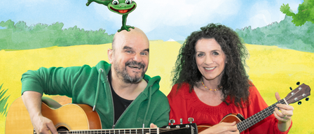Ein Mann und eine Frau sitzen in einem Feld und spielen beide Gitarre. Ein Frosch sitzt auf dem Kopf des Mannes. Sie lächeln beide für die Kamera.
