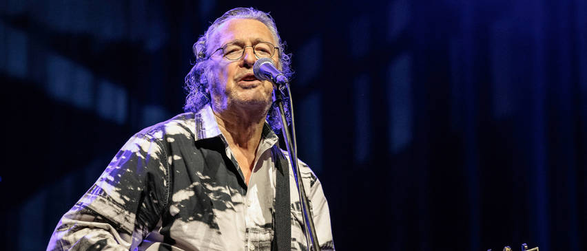 Ein Mann mit langen Haaren und Brille tritt auf der Bühne auf, spielt Gitarre und singt in ein Mikrofon.