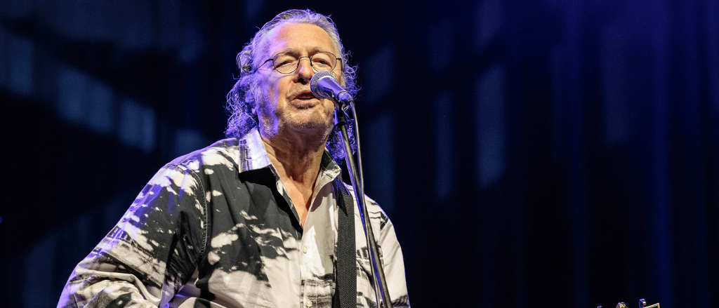 Ein Mann mit langen Haaren und Brille tritt auf der Bühne auf, spielt Gitarre und singt in ein Mikrofon.
