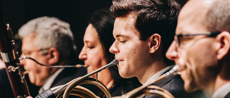 Eine Gruppe von Musikern spielt, wobei sie französische Hörner halten. Sie spielen in einem Konzert, und der Fokus liegt auf dem Mann in der Mitte.