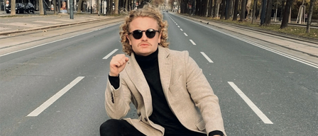 Ein Mann sitzt auf einem roten Mercedes Spielzeugauto in einer leeren Straße, trägt eine Sonnenbrille und ein Rollkragenshirt.