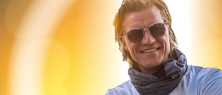 Ein Mann mit nassen Haaren und einem Lächeln trägt eine Sonnenbrille und ein weißes T-Shirt. Er hat einen grauen Schal um den Hals.