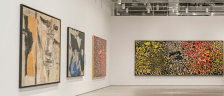 Eine Kunstgalerie mit weißen Wänden zeigt vier Gemälde. Das größte, farbenfrohe Gemälde befindet sich an der rechten Wand. Drei kleinere Gemälde hängen an der linken Wand.