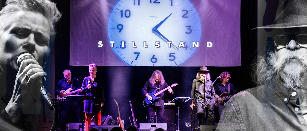 Eine Band tritt auf der Bühne auf, vor einem großen Hintergrund mit einer Uhr, die 11:10 anzeigt. Die Band besteht aus Gitarristen und einem Sänger. Das Publikum im Vordergrund ist verschwommen.