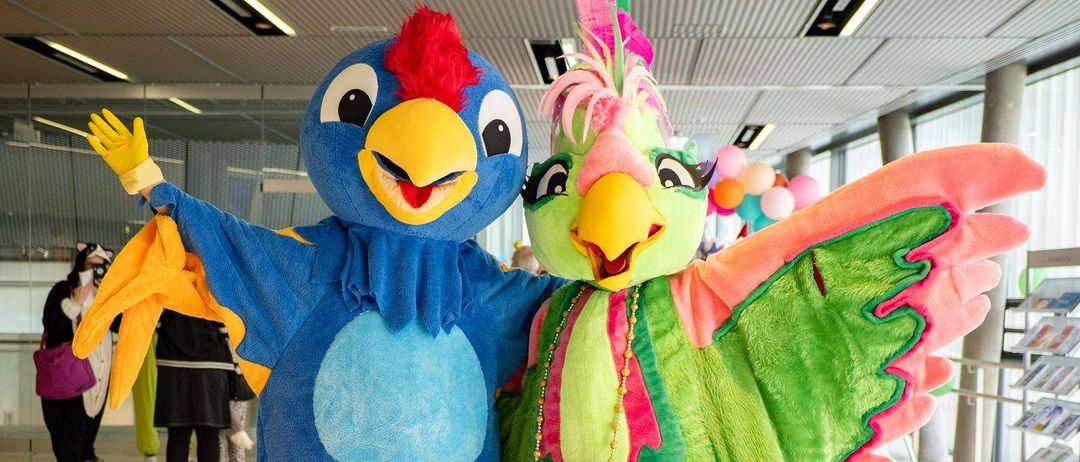 Zwei Menschen in farbenfrohen Papageikostümen stehen zusammen, einer mit einem blauen Körper und rotem Kamm, der andere mit einem grünen Körper und rosa Flügeln. Sie befinden sich in einem hell beleuchteten Innenraum mit Menschen und Luftballons im Hintergrund.