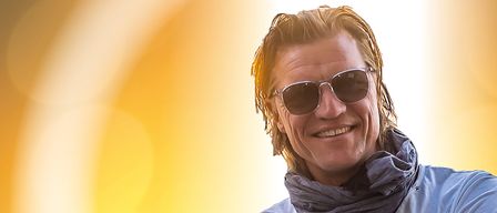 Ein Mann in einem weißen T-Shirt, einem grauen Schal und schwarzen Hosen posiert mit verschränkten Armen in einer sonnigen Umgebung.