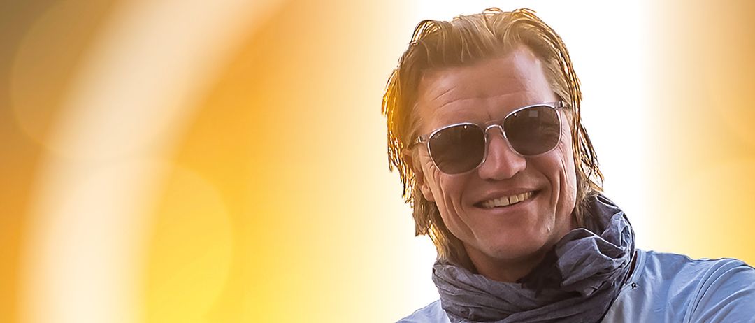 Ein Mann in einem weißen T-Shirt, einem grauen Schal und schwarzen Hosen posiert mit verschränkten Armen in einer sonnigen Umgebung.