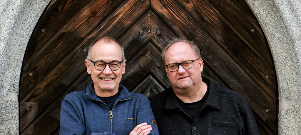 Zwei Männer stehen nebeneinander vor einer Holztür und lächeln beide, beide tragen eine Brille.
