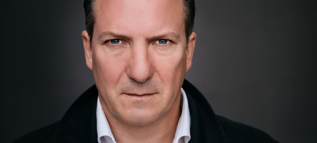Ein Porträt eines Mannes mittleren Alters mit blauen Augen, der ein weißes Hemd und eine schwarze Jacke vor grauem Hintergrund trägt.