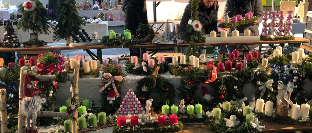 Ein Weihnachtsmarkt zeigt Kränze, Kerzen und Dekorationen, die auf einem Tisch arrangiert werden. Im Hintergrund sind weitere festliche Artikel ausgestellt, und es sind Menschen beim Stöbern zu sehen.