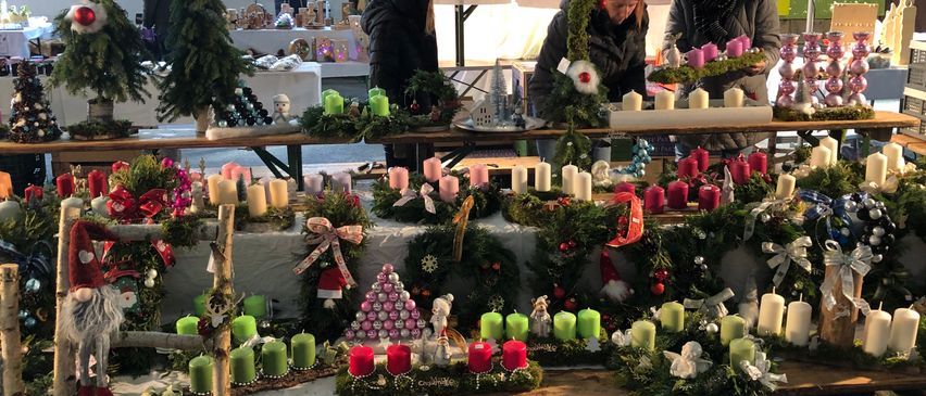 Ein Weihnachtsmarkt zeigt Kränze, Kerzen und Dekorationen, die auf einem Tisch arrangiert werden. Im Hintergrund sind weitere festliche Artikel ausgestellt, und es sind Menschen beim Stöbern zu sehen.