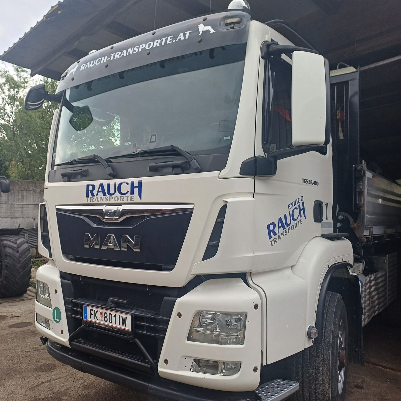 Ein weißer MAN-Lkw mit 'RAUCH TRANSPORTE' auf der Front, geparkt in einer Garage mit einer Ziegelmauer im Hintergrund.