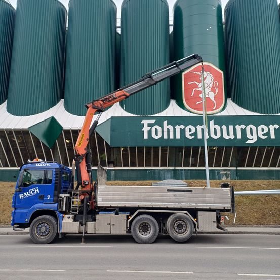 Ein blauer Lastwagen mit einem Kran steht vor dem Fohrenburger-Gebäude mit großen grünen Silos.