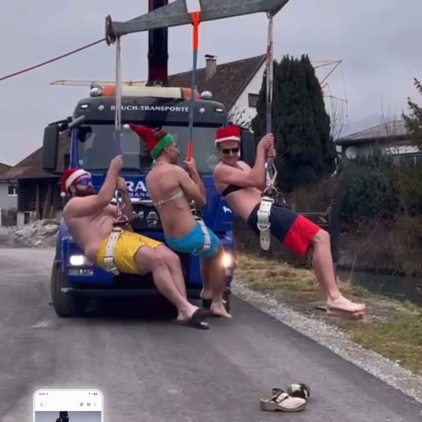 Drei Männer in Santa-Hüten und Badeshorts hängen an einem LKW, wahrscheinlich als weihnachtlicher Scherz. Der LKW trägt den Aufdruck 'Ruch-Transporte'.
