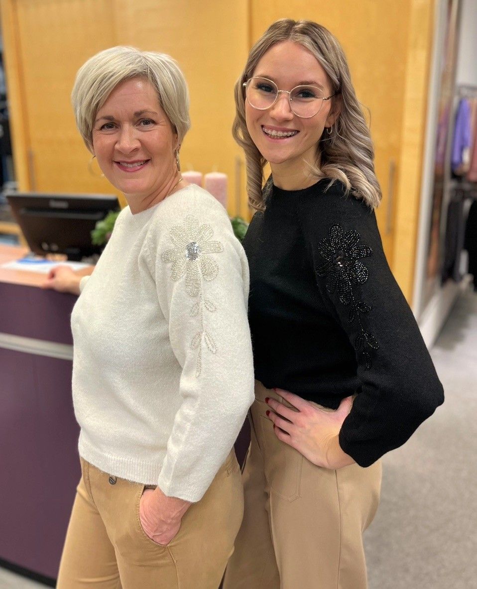 Zwei Frauen stehen nebeneinander in einem Bekleidungsgeschäft. Die Frau links trägt einen weißen Pullover mit Blumendesign, während die Frau rechts einen schwarzen Pullover mit Blumendesign trägt. Sie lächeln beide und scheinen für ein Foto zu posieren. Hinter ihnen steht ein Schreibtisch mit Monitor und Kerzen.