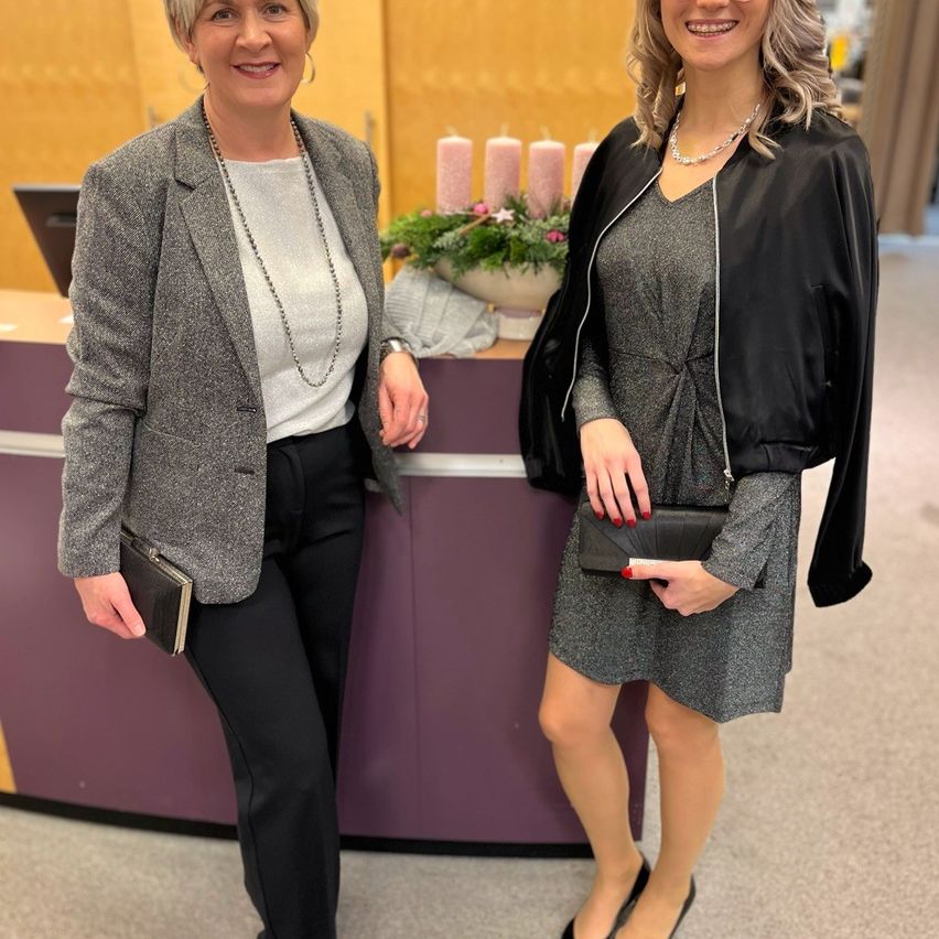 Zwei Frauen stehen neben einem Empfangstresen mit einer Blumenarrangement und lächeln in die Kamera. Eine hält ein Buch und eine Handtasche, die andere eine Clutch.
