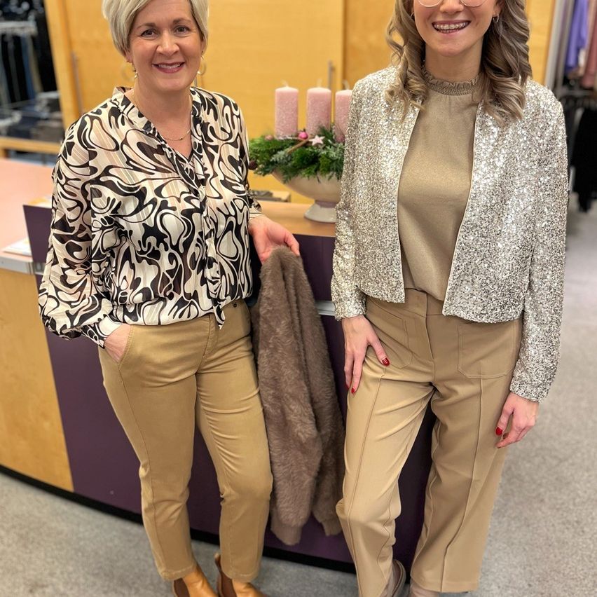 Zwei Frauen stehen in einem Bekleidungsgeschäft, beide tragen beige Hosen. Eine Frau hat kurze blonde Haare, die andere hat lange blonde Haare und trägt eine Paillettenjacke. Hinter ihnen steht eine Theke mit einer Pflanze und Kerzen.