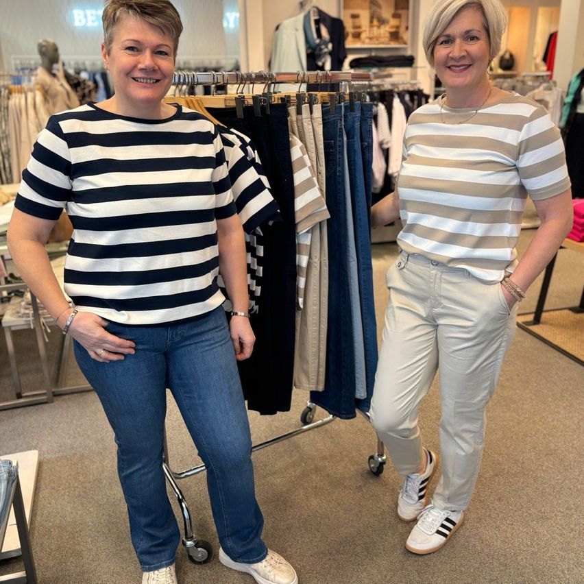 Zwei Frauen stehen in einem Bekleidungsgeschäft, eine mit einem gestreiften Shirt und die andere mit Hosen. Sie lächeln und scheinen auf Kleidung an einem Kleiderständer hinter ihnen zu schauen.