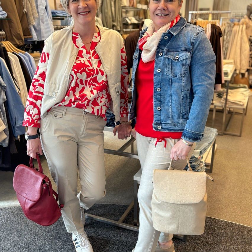 Zwei Frauen stehen in einem Bekleidungsgeschäft, beide halten Taschen. Eine trägt ein rotes Blumenhemd und beige Hosen, die andere ein rotes Oberteil und eine Jeansjacke mit einem Schal. Hinter ihnen hängen Kleidungsstücke an Stangen.