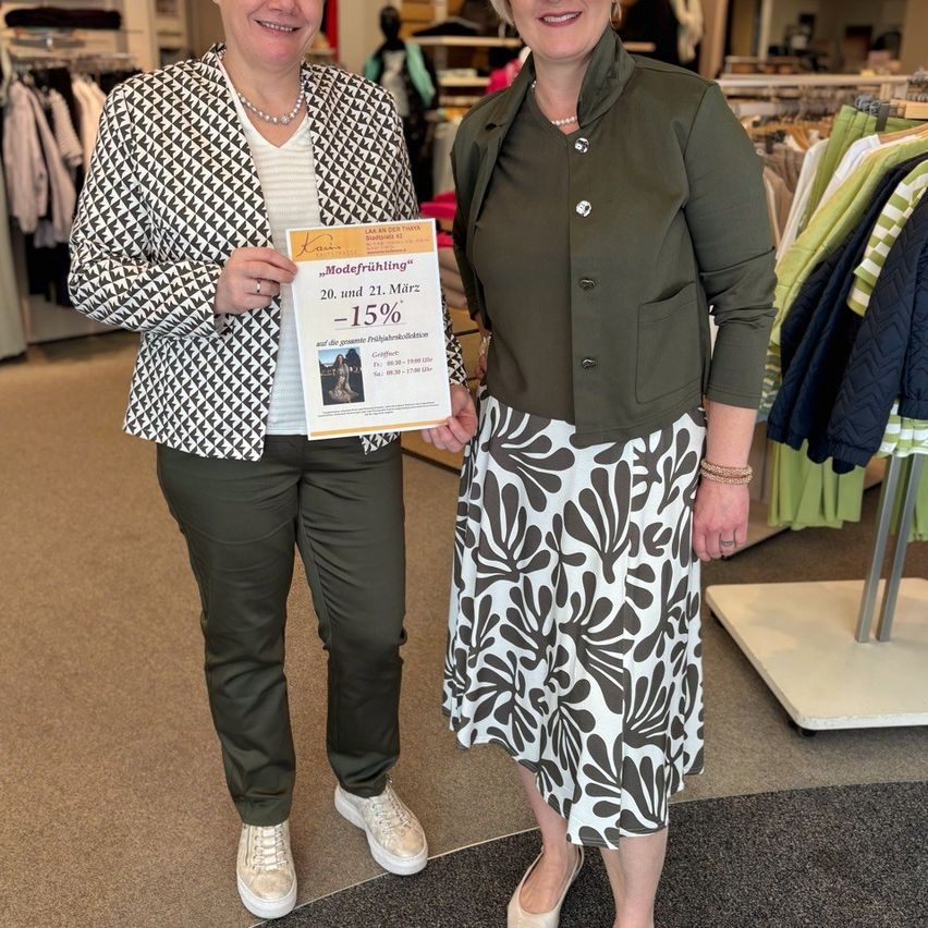 Zwei Frauen stehen in einem Bekleidungsgeschäft, eine hält ein Schild mit der Aufschrift 'Modefrühling' und einem 15% Rabatt vor Kleiderstangen.