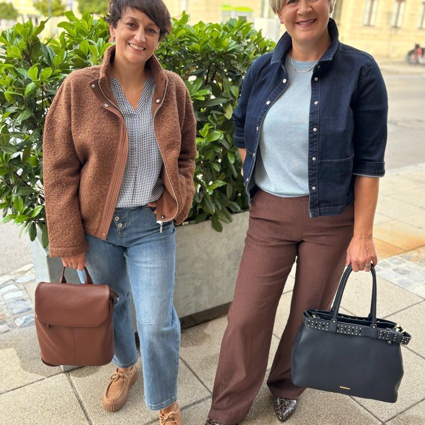 Zwei Frauen stehen auf einem Bürgersteig, eine hält eine braune Ledertasche und die andere eine schwarze Lederhandtasche mit Nieten, beide lächeln und tragen lässige Outfits.