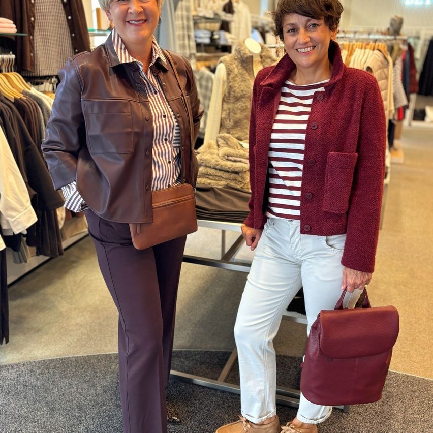 Zwei Frauen stehen in einem Bekleidungsgeschäft, eine trägt eine braune Lederjacke und die andere einen roten Cardigan mit weißen Hosen. Sie lächeln beide und halten Taschen.