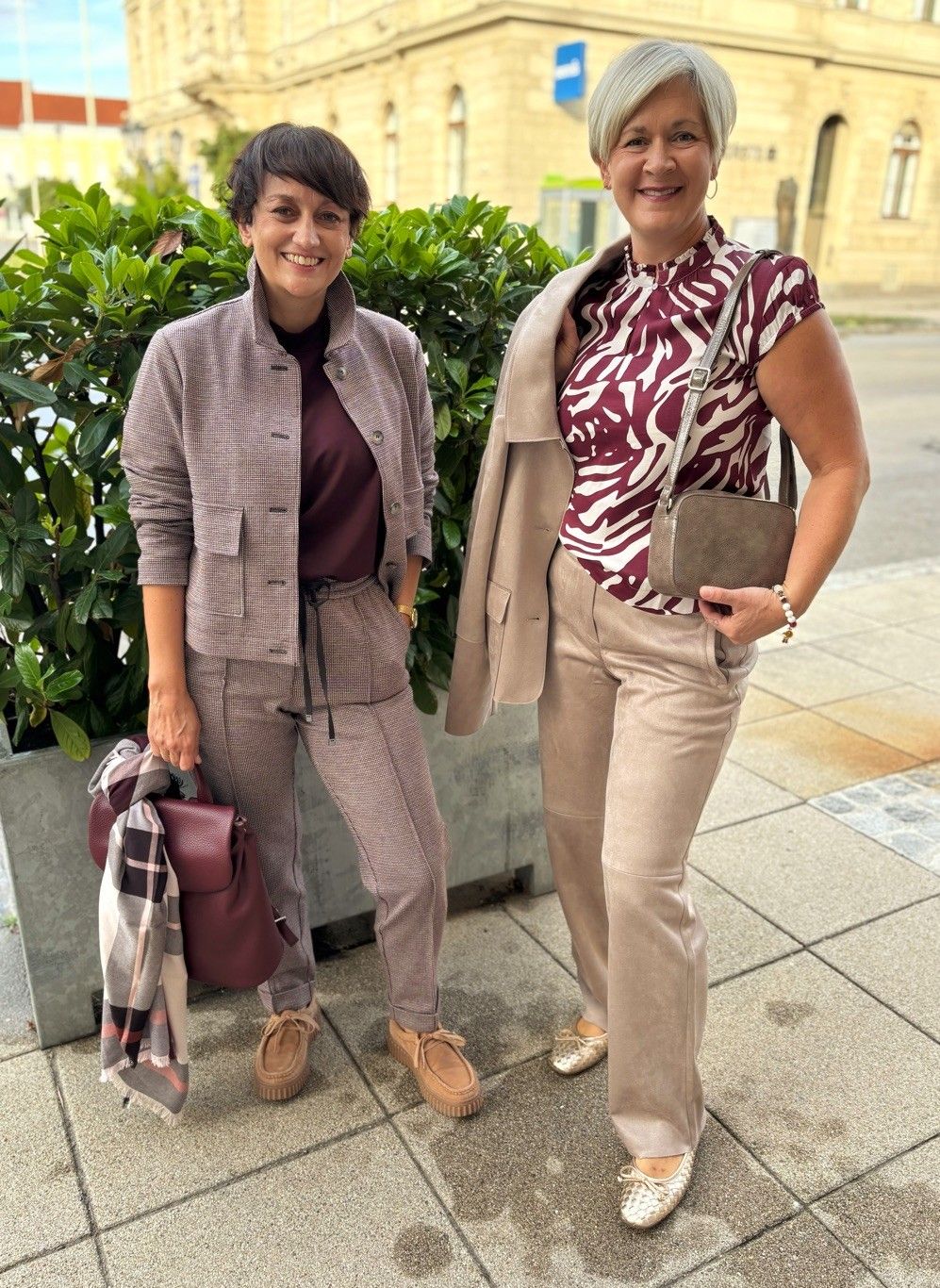 Zwei Frauen in herbstlichen Outfits stehen auf einem Bürgersteig, eine hält eine rote Handtasche und die andere trägt einen karierten Schal.
