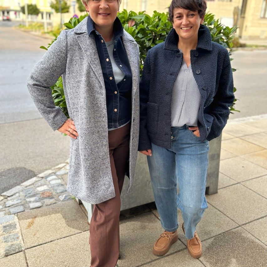 Zwei Frauen stehen auf einem Bürgersteig, eine in einem grauen Mantel und die andere in einer blauen Jacke, beide lächeln. Hinter ihnen steht eine große grüne Pflanze in einem Pflanzgefäß.