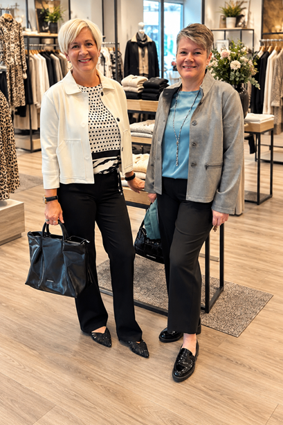 Two women stand in a clothing store, smiling and posing for a photo. One holds a black handbag, and both wear black pants and jackets. Behind them, clothing racks and a table with flowers are visible.