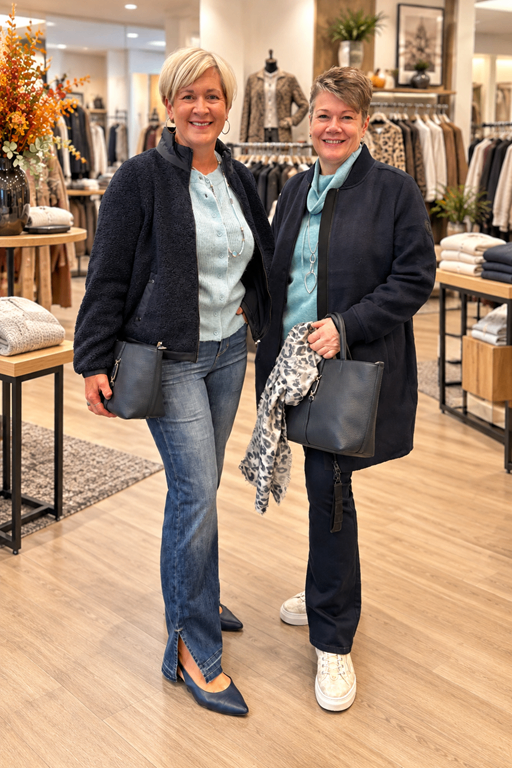 Two women stand in a clothing store, smiling and holding handbags. One wears a blue jacket and jeans, the other a black coat with a scarf.
