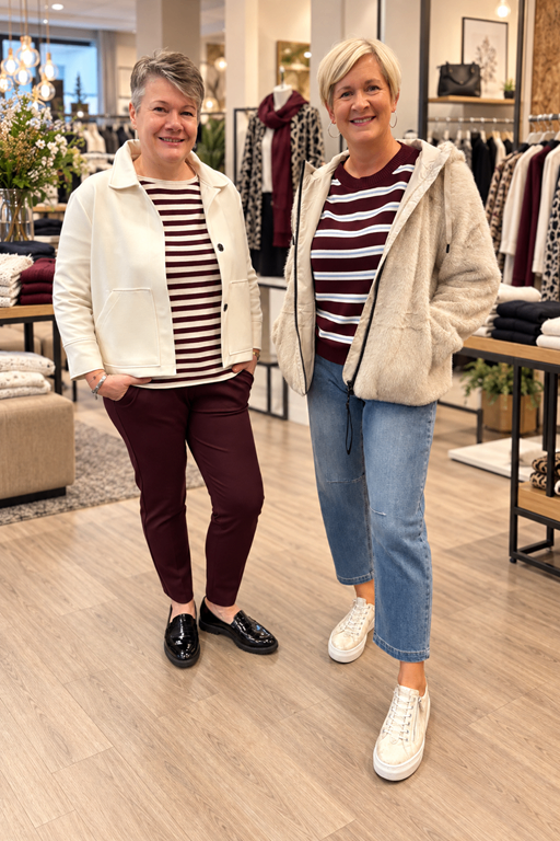Two women are standing in a clothing store, smiling and posing for a photo. One is wearing a white jacket and maroon pants, the other a beige jacket and jeans.