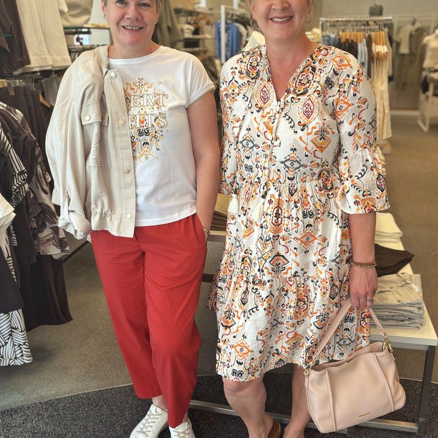 Zwei Frauen stehen in einem Bekleidungsgeschäft. Eine trägt ein weißes T-Shirt und rote Hosen, die andere ein gemustertes Kleid mit einer beigefarbenen Tasche. Sie lächeln für die Kamera.
