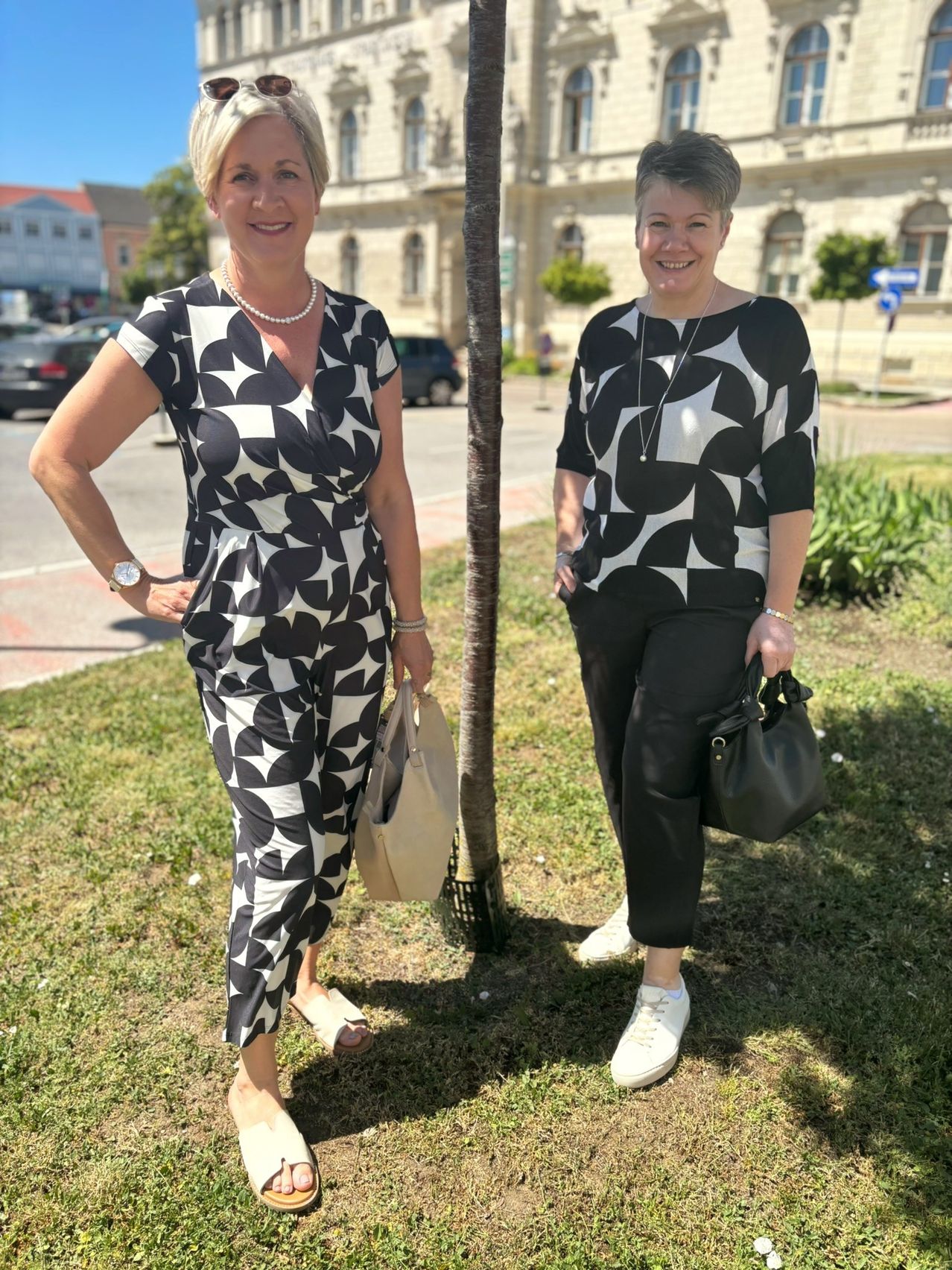 Zwei Frauen stehen neben einem Baum auf einem Rasen. Beide lächeln und sind in Schwarz und Weiß gekleidet. Sie halten Handtaschen und stehen vor einer Stadtstraße mit geparkten Autos.