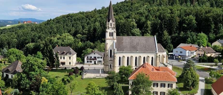 Luftaufnahme einer Kirche mit einem Turm, umgeben von Häusern und Bäumen. Die Kirche befindet sich in einem grünen Bereich mit klarem blauen Himmel.