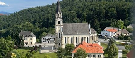 An aerial view of a church surrounded by houses and greenery, set against a backdrop of hills and trees.
